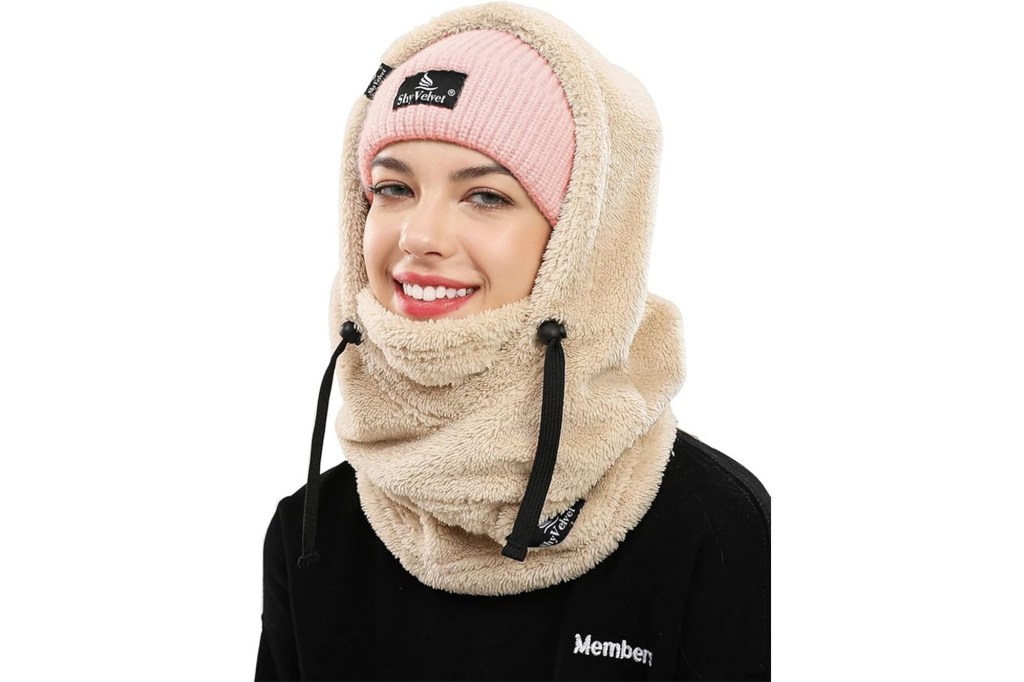 A woman wearing a pink knitted beanie with a "Shy Velvet" tag, covered by a beige faux fur hood and matching neck gaiter with drawstrings.