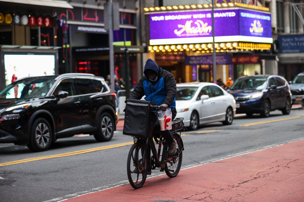 A masked food delivery worker on a bicycle with a large delivery bag on the handlebars and two drinks hanging from the side of the bike, rides past cars and in front of an "Aladdin" sign in Times Square.