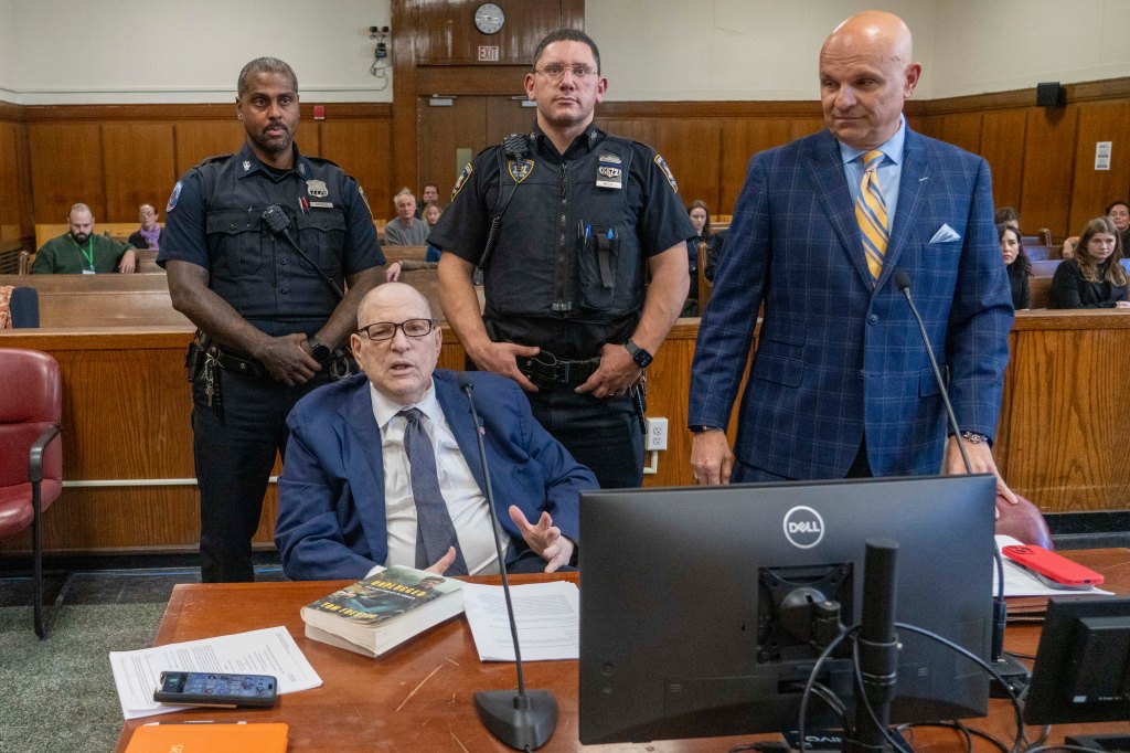 Harvey Weinstein in court, seated at a table with a book, two police officers standing behind him, and a man in a blue plaid suit and yellow tie to his right.