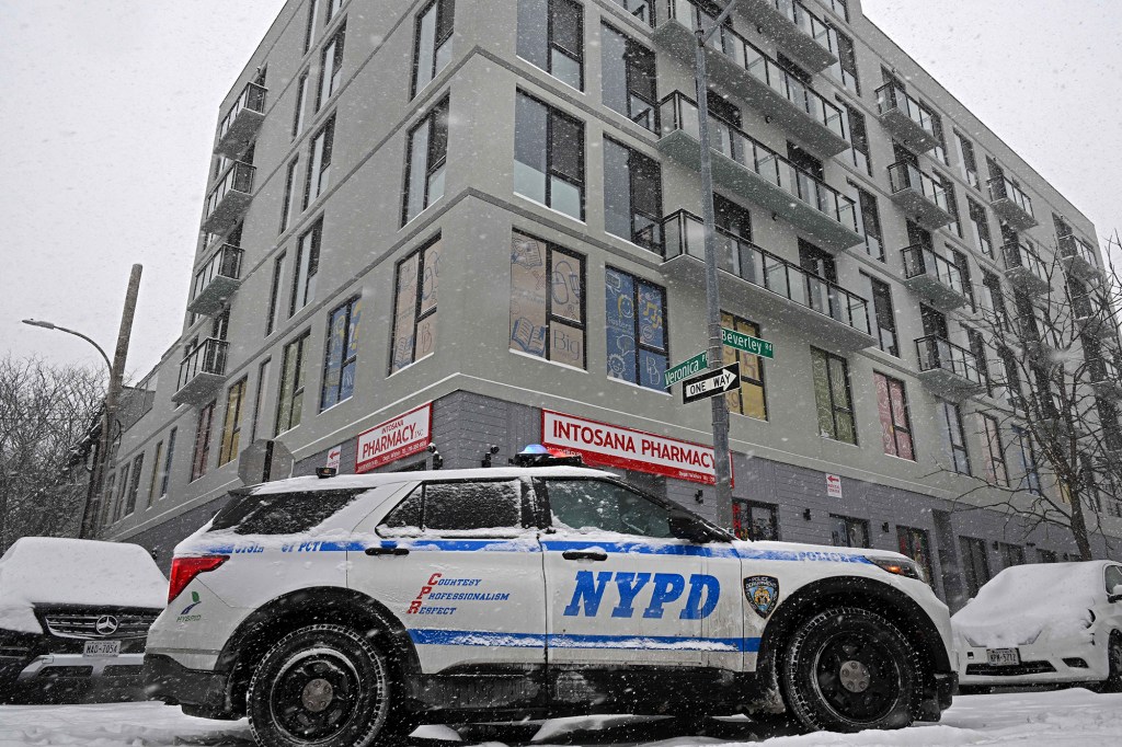 An NYPD police car parked on a snow-covered street next to a building with a pharmacy on the ground floor.