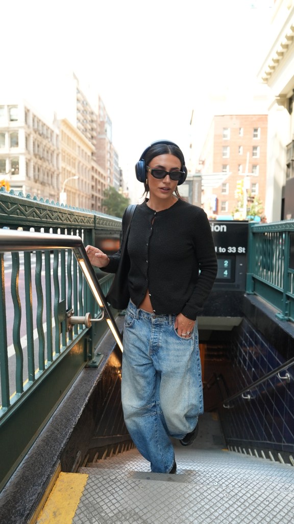 Musician Fia James descending subway steps, wearing headphones, sunglasses, a black cardigan, and baggy jeans.