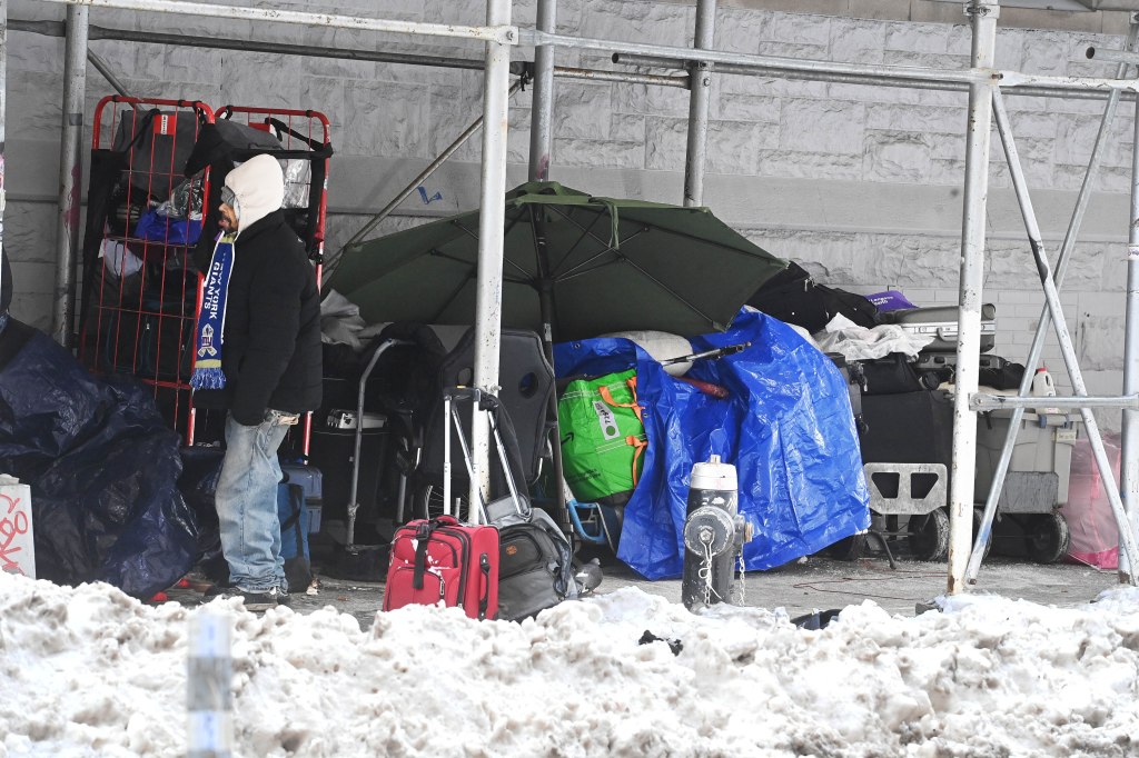 A homeless encampment under scaffolding with a person wearing a New York Giants scarf standing in snow.