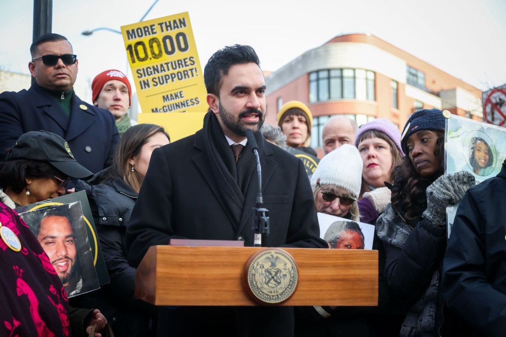 Mayor Mamdani speaks at a podium with a crowd holding portraits and signs that read "More than 10,000 Signatures in Support! Make McGuinness" behind him.