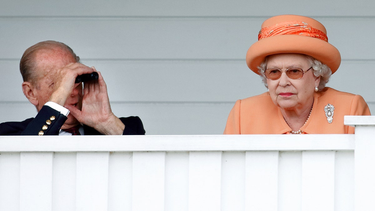Queen Elizabeth looks sternly in an orange coatdress while Prince Philip looks on from his binoculars.