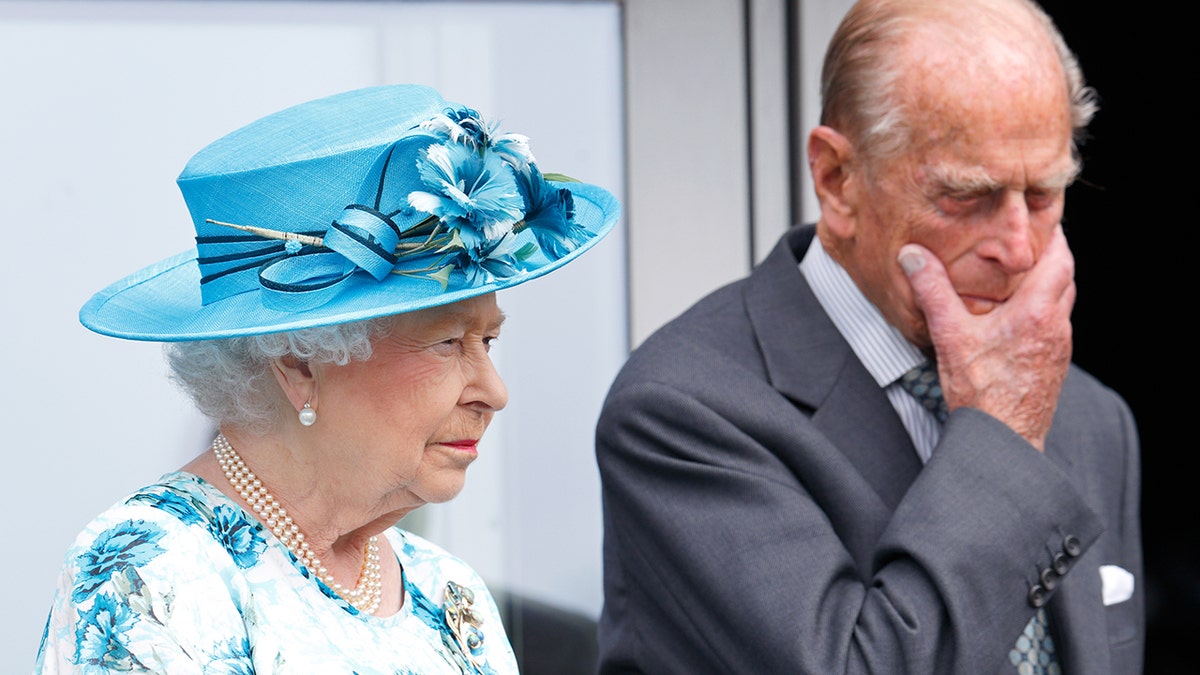 Prince Philip with his hand to his mouth as a stern Queen Elizabeth looks on during a royal engagement.