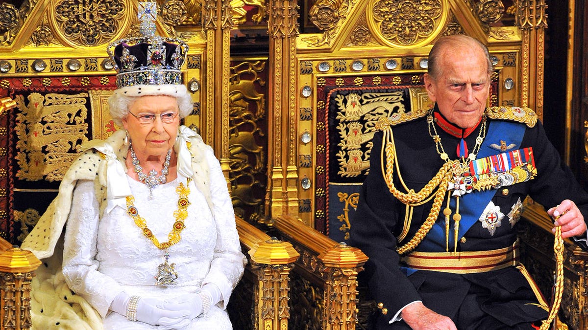 Queen Elizabeth wearing a white gown and a crown sitting next to Prince Philip in royal regalia.