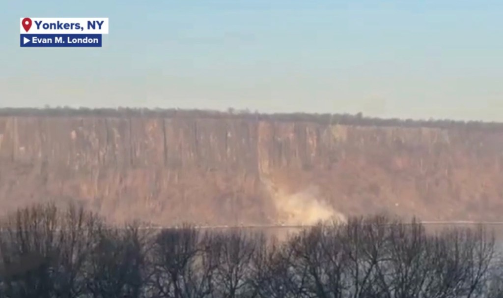 A rockslide on a cliff overlooking a body of water, with a cloud of dust rising from the base of the cliff.