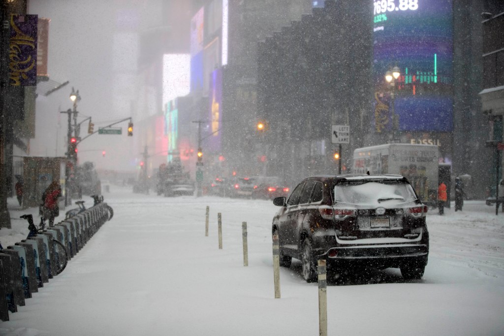 A snow-covered street in New York City during a snowstorm, with Times Square billboards and a parked SUV in the foreground.