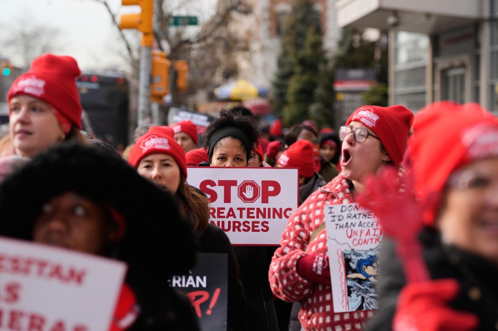 Striking nurses in red hats and winter coats, one with a sign reading "STOP THREATENING NURSES."