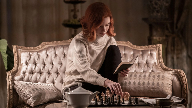 Beth Harmon reading a book while wearing a white sweater and sitting on a couch in front of a chess set in The Queen's Gambit