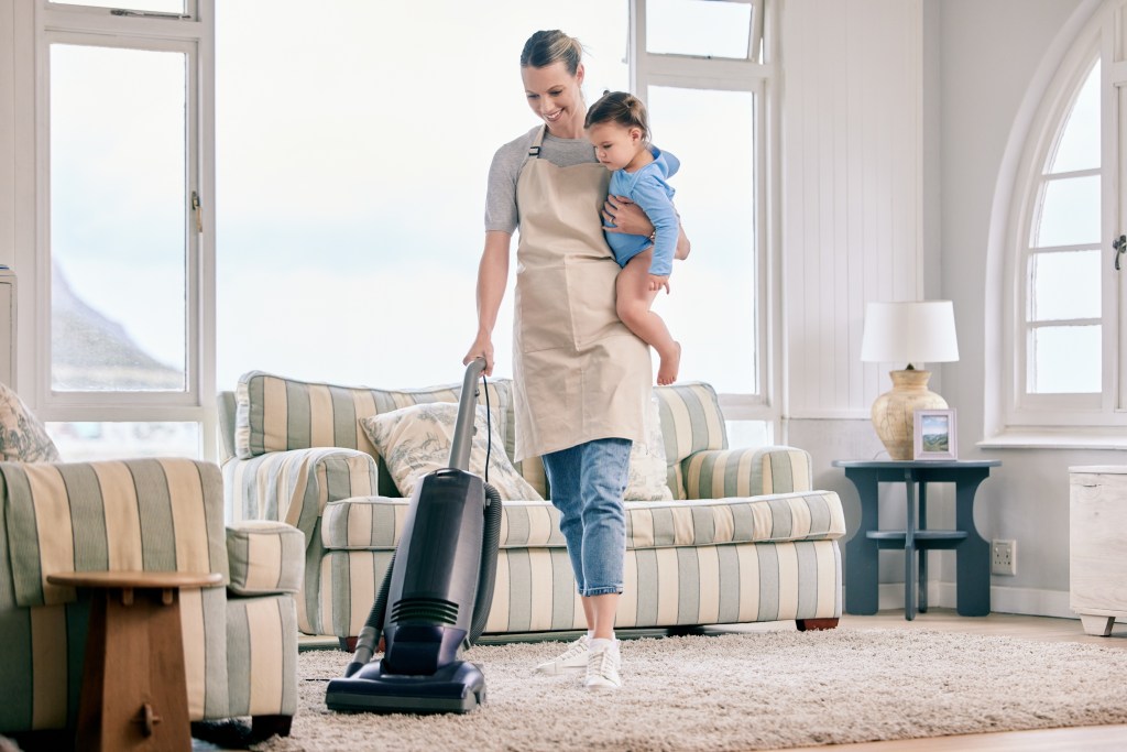 A mother holding her toddler while vacuuming the living room carpet.