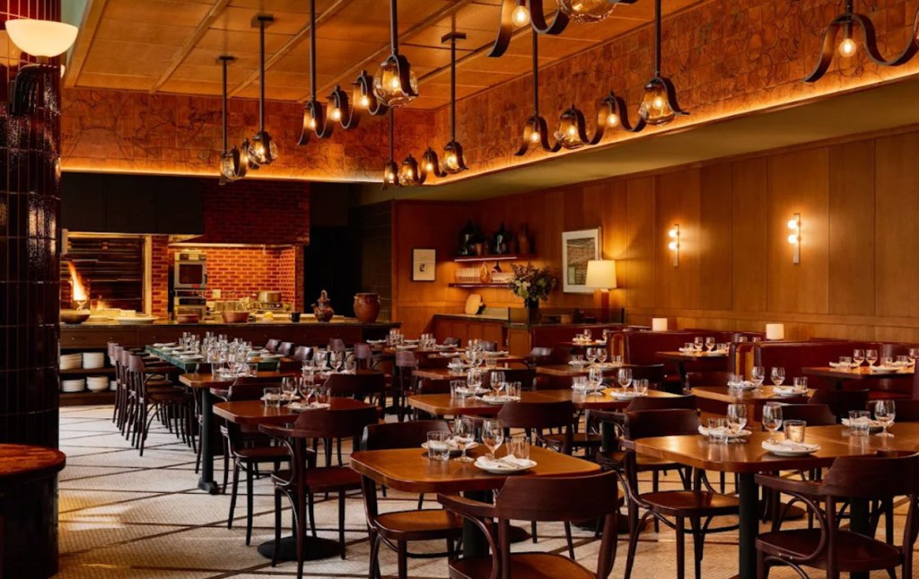 A restaurant interior with wooden tables and chairs set with glasses and plates, an open kitchen with a brick wall and a wood-fired oven, and warm lighting from hanging fixtures and wall sconces.