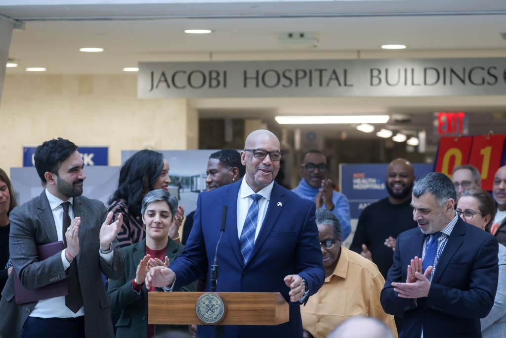 Stanley Richards speaks at a podium as Zohran Mamdami and other people clap.
