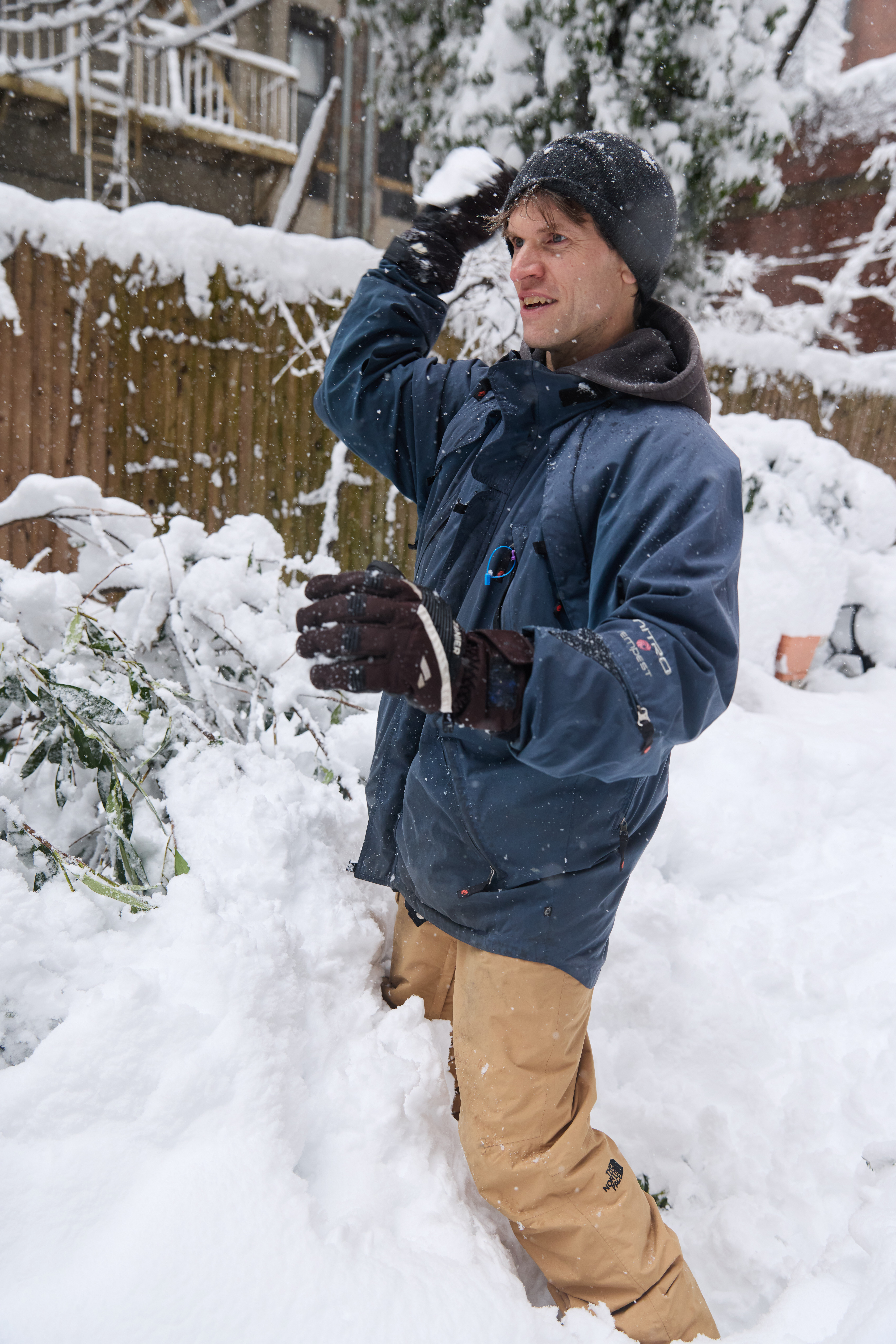 Will Pavia playing in the snow, wearing a blue jacket, black gloves, and a black hat, surrounded by snow-covered bushes and a wooden fence.