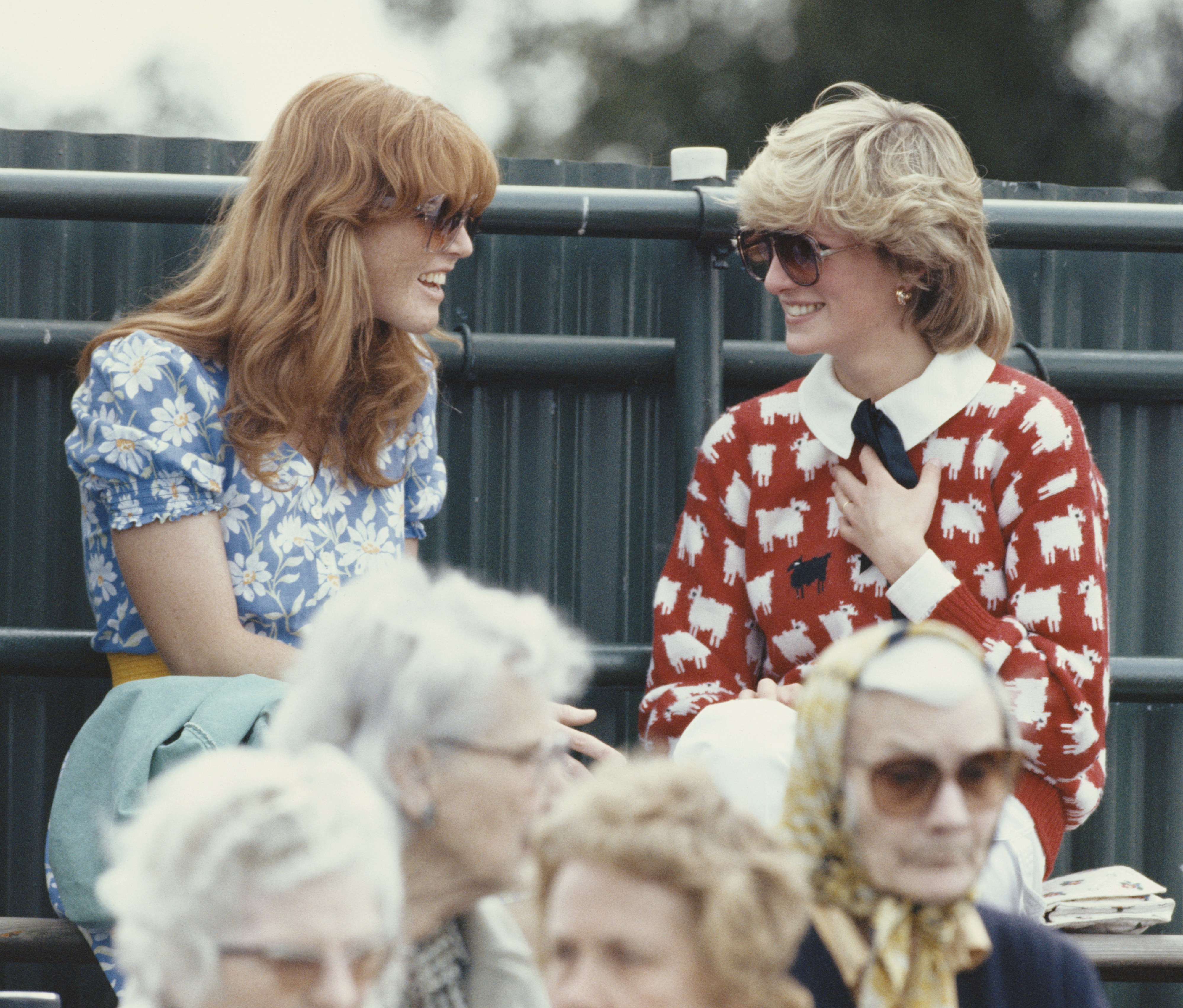 Princess Diana in a red sheep-patterned sweater and Sarah Ferguson in a blue floral dress, both wearing sunglasses, laughing in a crowd.