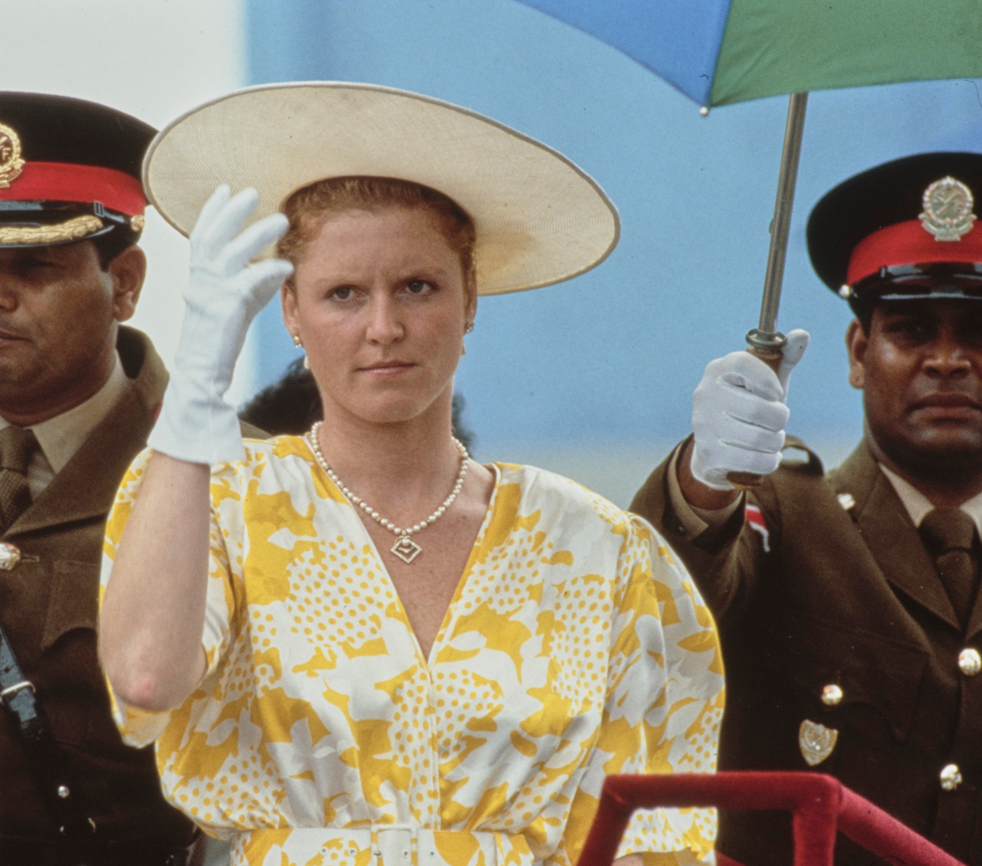 Sarah, Duchess of York, in a yellow-and-white dress and wide-brim hat, beside Prince Andrew, Duke of York, in a Royal Navy uniform, with military figures.