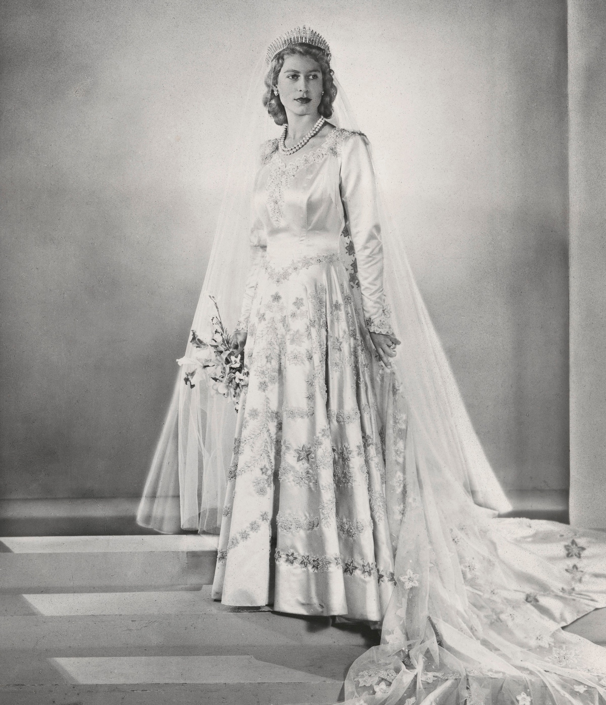 A full-length black and white photo of Queen Elizabeth II in her wedding dress, tiara, veil, and holding a bouquet, standing on stairs.