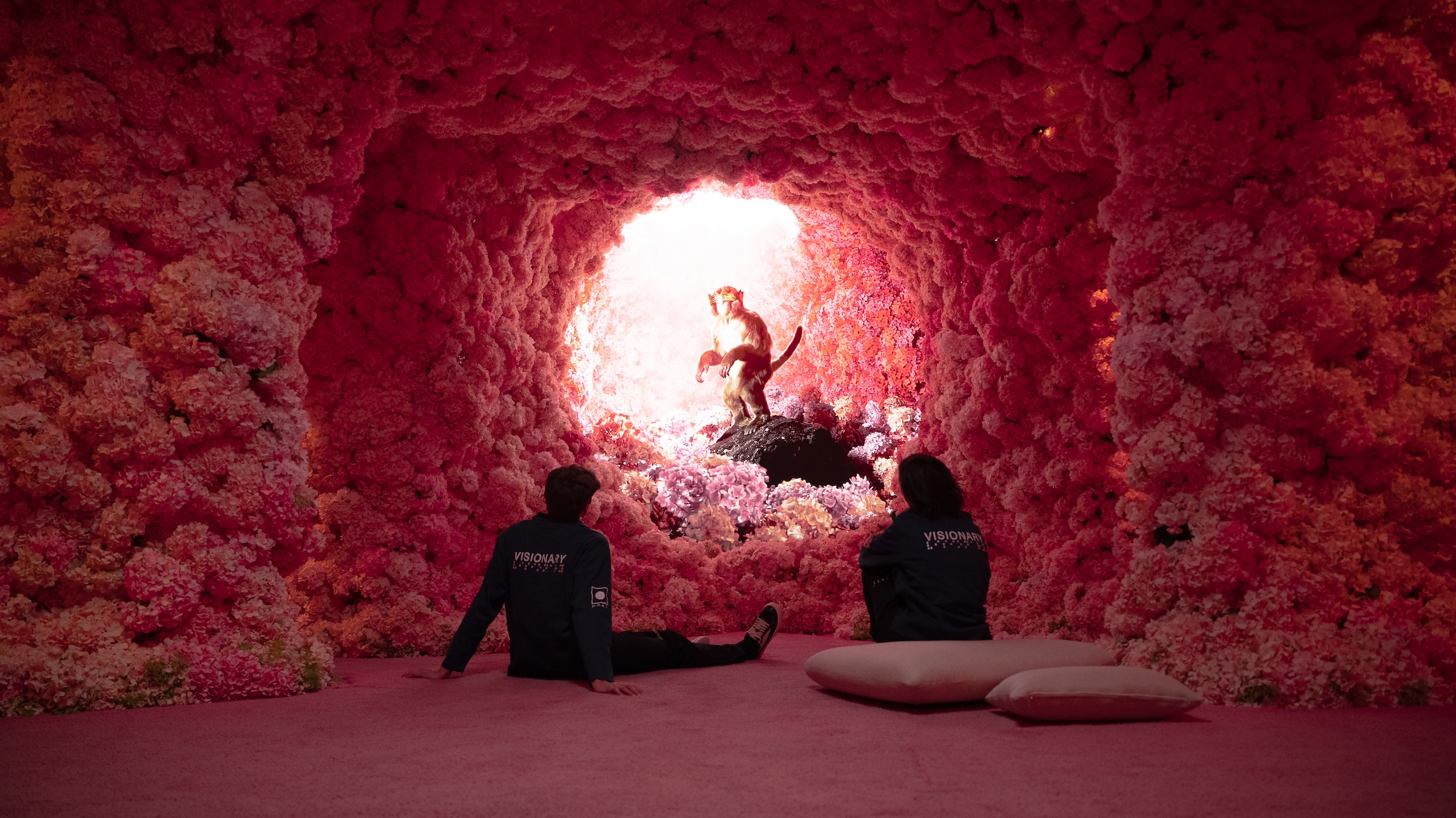 Two people sit on a pink carpeted floor, viewed from behind, inside a cave-like room completely covered in pink and white hydrangeas. They are looking toward a bright, circular opening at the end of the floral tunnel. In the center of the light stands a life-sized golden monkey statue atop a dark rock. The individuals are wearing dark blue jackets with the words "VISIONARY LABS" printed on the back. Two large pink pillows rest on the floor to the right.