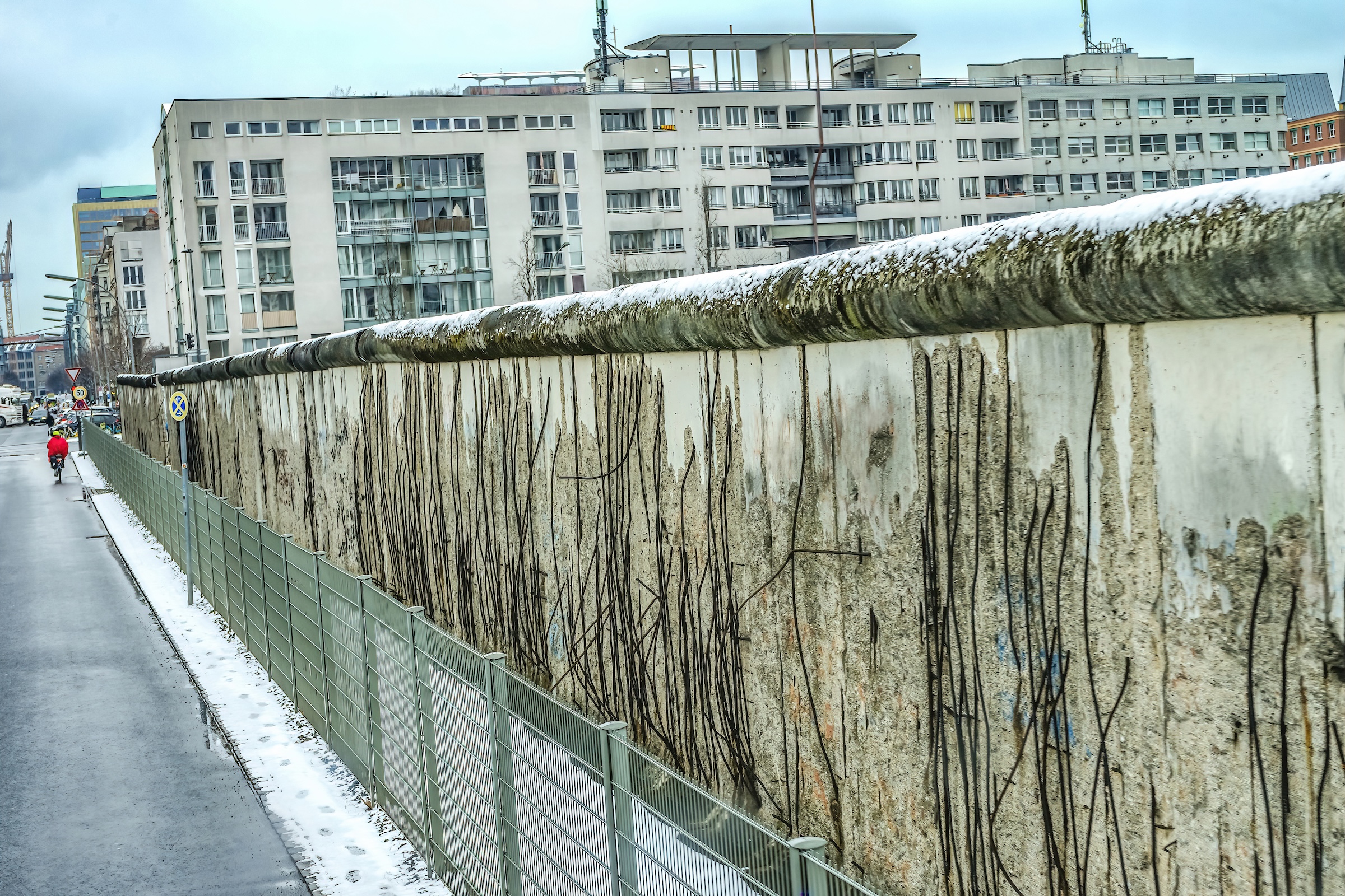 A long, weathered section of the Berlin Wall stands under a cloudy sky with a light dusting of snow on its rounded top. The concrete surface is heavily eroded, exposing vertical rusted iron rebar throughout. A green metal fence runs parallel to the wall on a damp asphalt path, with modern apartment buildings visible in the background.