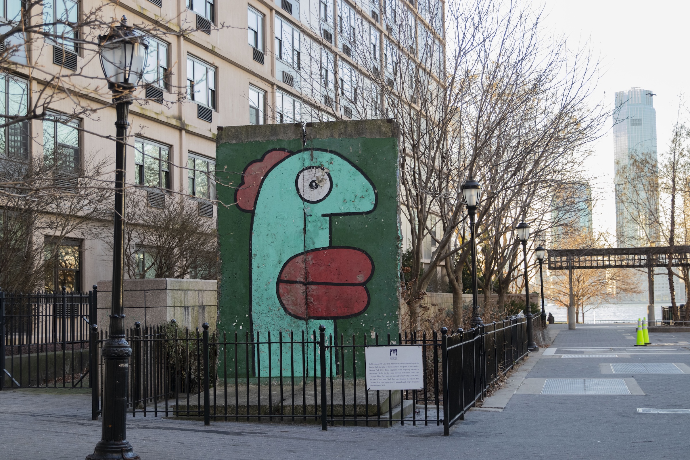 A wide shot of the Berlin Wall segment located in Kowsky Plaza, New York City. The colorful graffiti-covered slab sits behind a black fence, flanked by traditional black lamp posts and bare winter trees. In the background, modern city buildings and a glimpse of the Hudson River are visible under a clear, bright sky.
