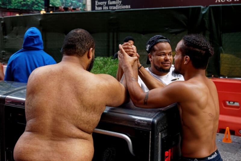 Two shirtless men arm wrestle on a trash bin outdoors as a third man in a white shirt and cap watches closely. A person in a blue hoodie stands in the background near a green fence and an orange barrier.
