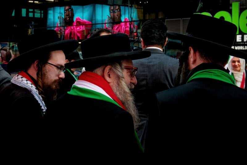 Three men in traditional Jewish attire and hats stand together, wearing scarves in the colors of the Palestinian flag. Bright billboards and other people are visible in the background.