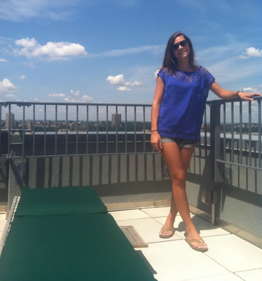 A woman in sunglasses stands on a rooftop patio, leaning on a railing with a clear sky in the background