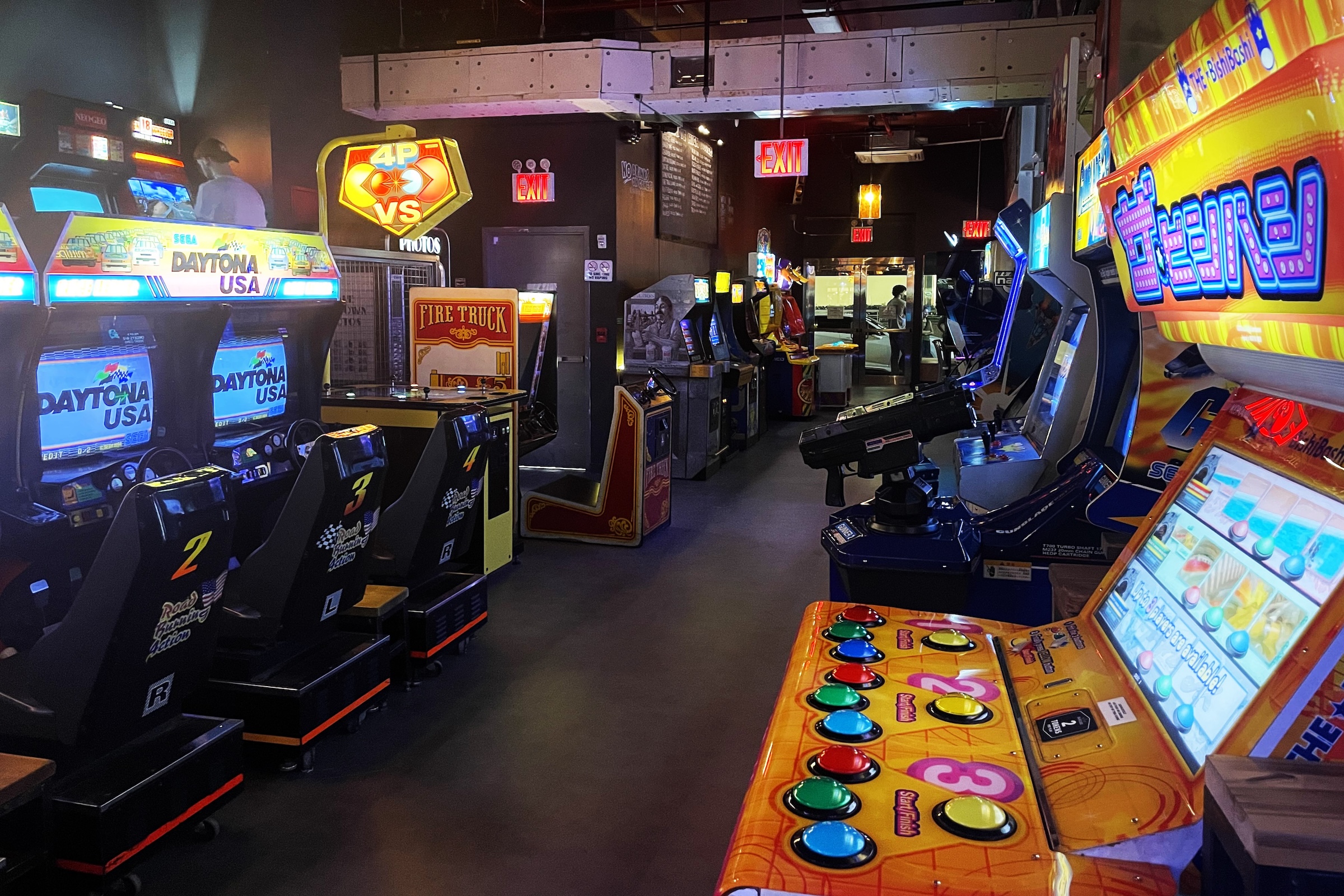 A row of classic arcade racing and shooting games, including multiple Daytona USA driving simulators, a Fire Truck game, and colorful button-based machines, inside a dimly lit arcade.
