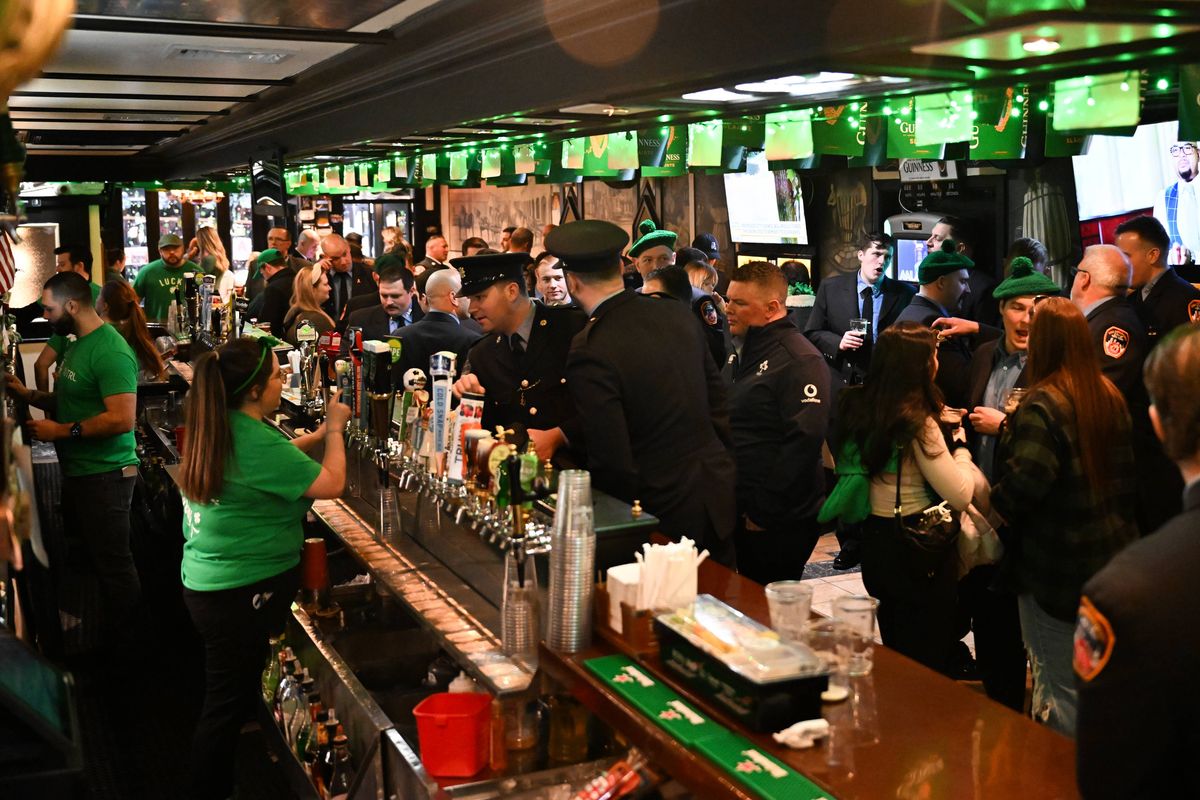 Bar staff pour pints while wearing green t-shirts to celebrate St Patrick's Day. The dimly lit pub is full of customers and there is green bunting hanging from the ceiling. 