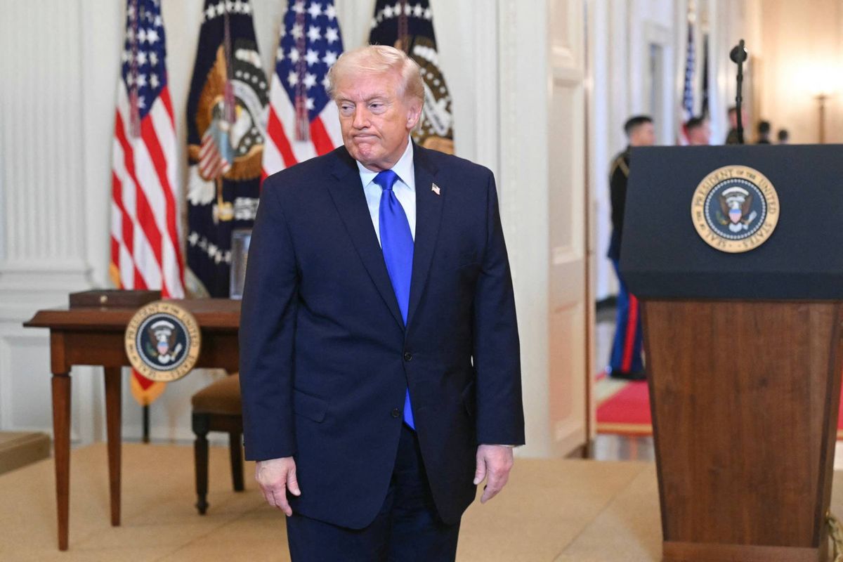 A man in a formal suit and tie is standing in an office setting, with a podium and flags in the background.