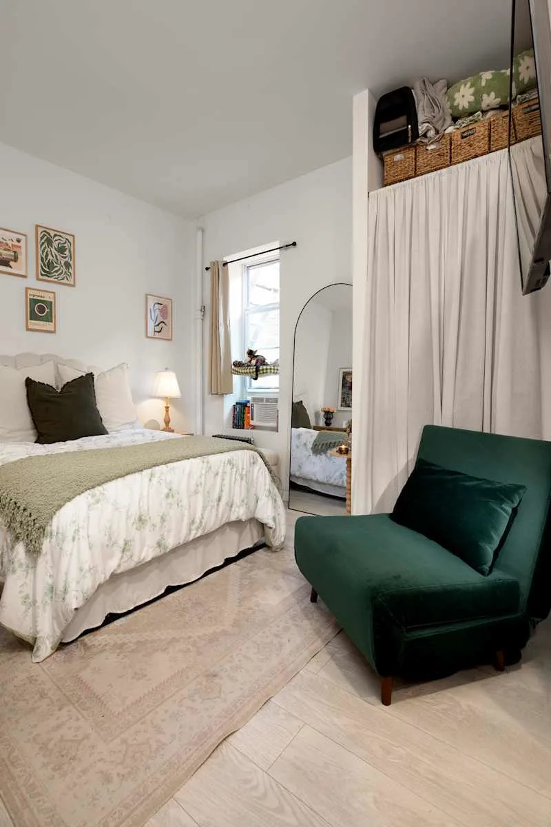 Cozy bedroom featuring a bed with floral bedding, a green velvet chair, and a large mirror. Decor includes framed art and a window.