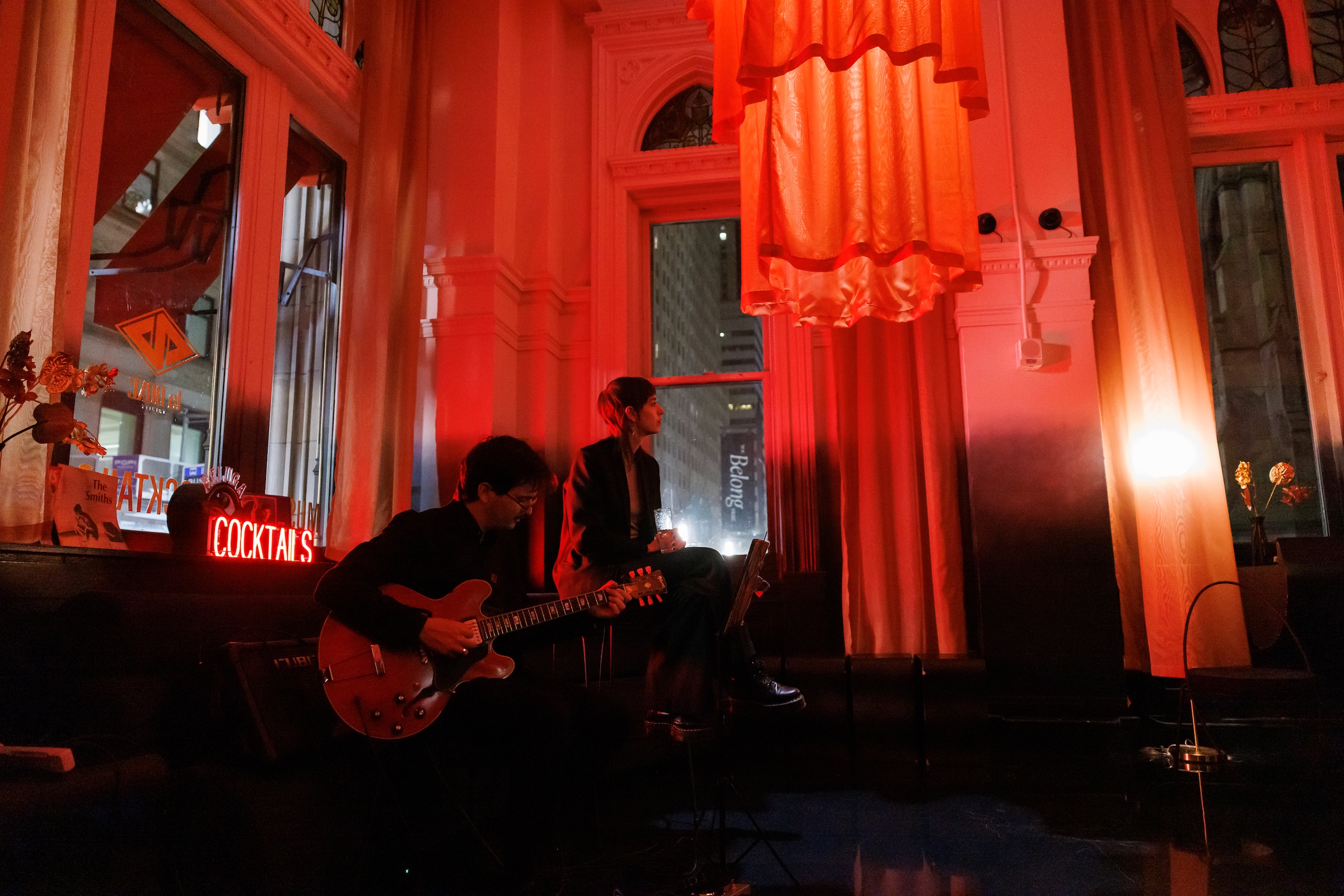 A dimly lit lounge bathed in red light, where a guitarist performs beside a person seated with a drink. The space features tall arched windows, draped curtains, and a glowing red “COCKTAILS” sign, creating a moody, intimate atmosphere.