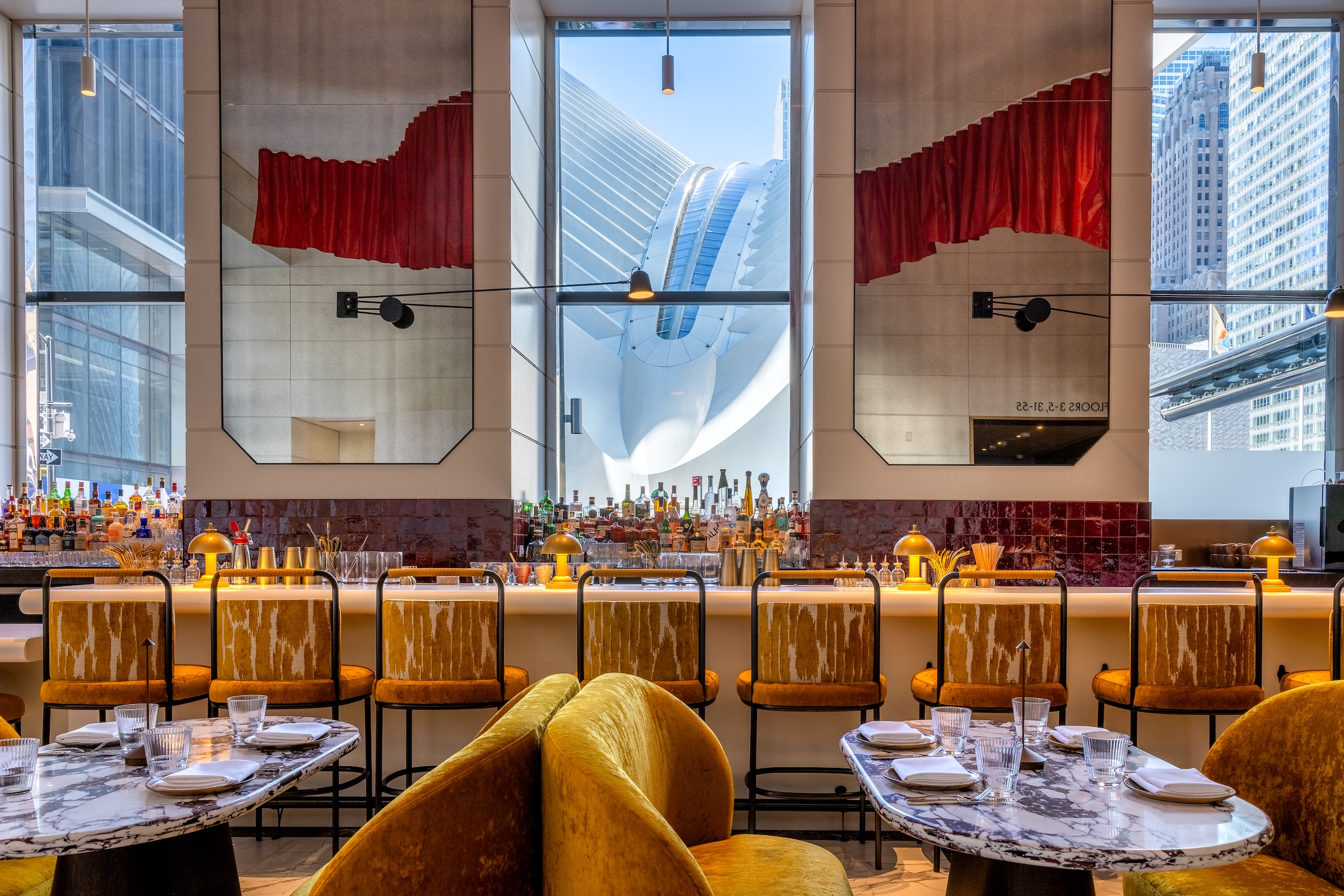 A stylish restaurant interior with gold velvet seating, marble tables, and a long bar lined with matching gold-upholstered barstools. Behind the bar, shelves of liquor bottles sit in front of tall windows. Through the center window, the white, wing-like structure of the Oculus at the World Trade Center is prominently visible. Large decorative mirrors with red curtain–like designs hang on either side, reflecting the modern space.