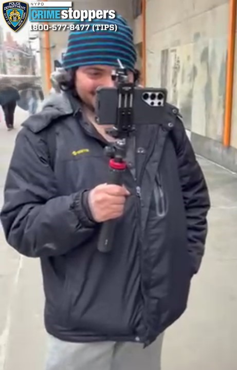 A man with a light complexion wearing a black jacket, light-colored pants, and a multicolored hat, holding a phone on a stick.