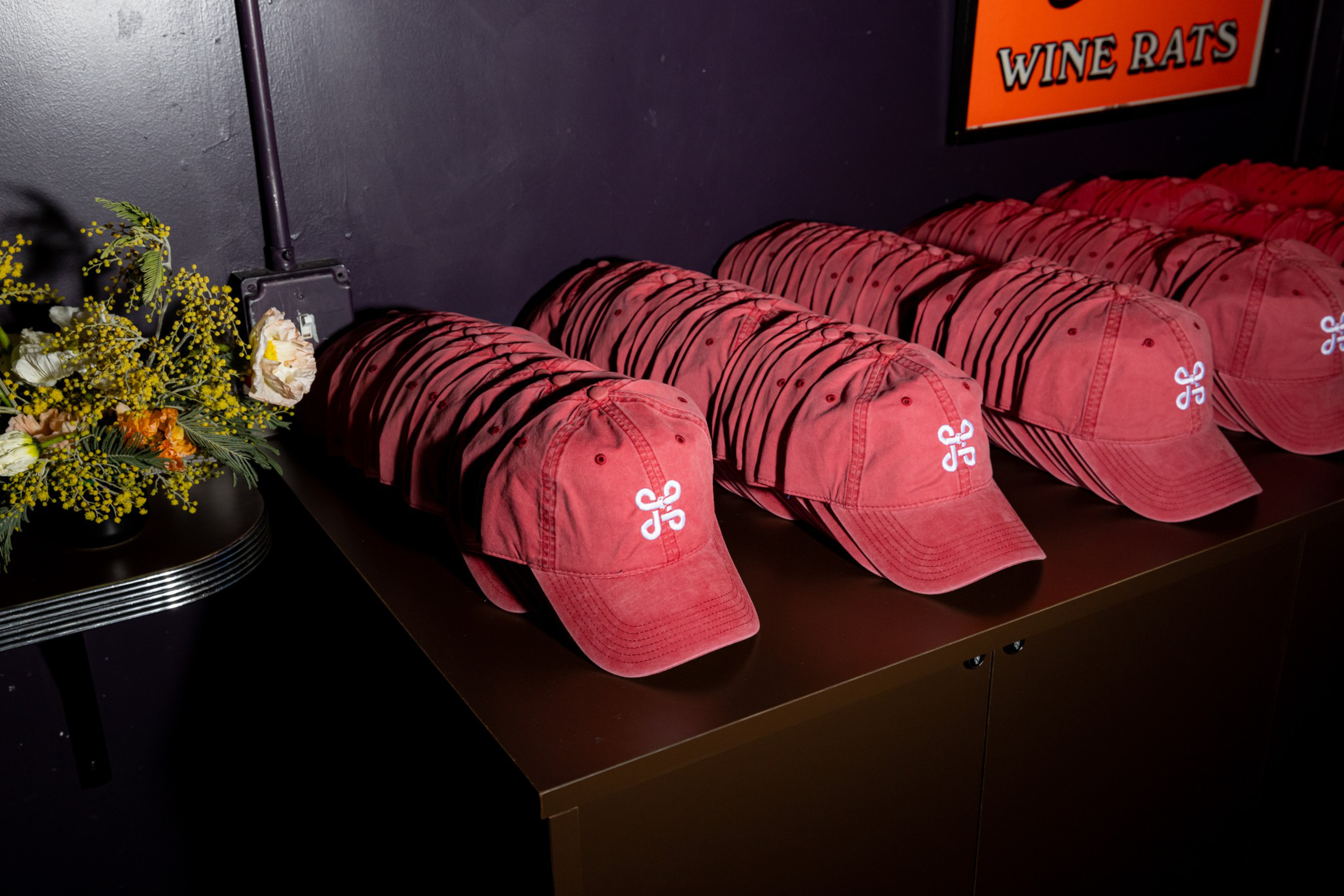Multiple red baseball caps with a white embroidered symbol are neatly lined up on a dark shelf, next to a bouquet of yellow and white flowers.