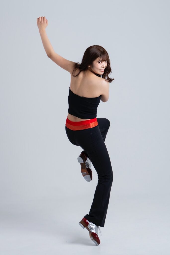 A person wearing a black sleeveless top and black pants with a red waistband, jumping while dressed in tap shoes, set against a plain background.