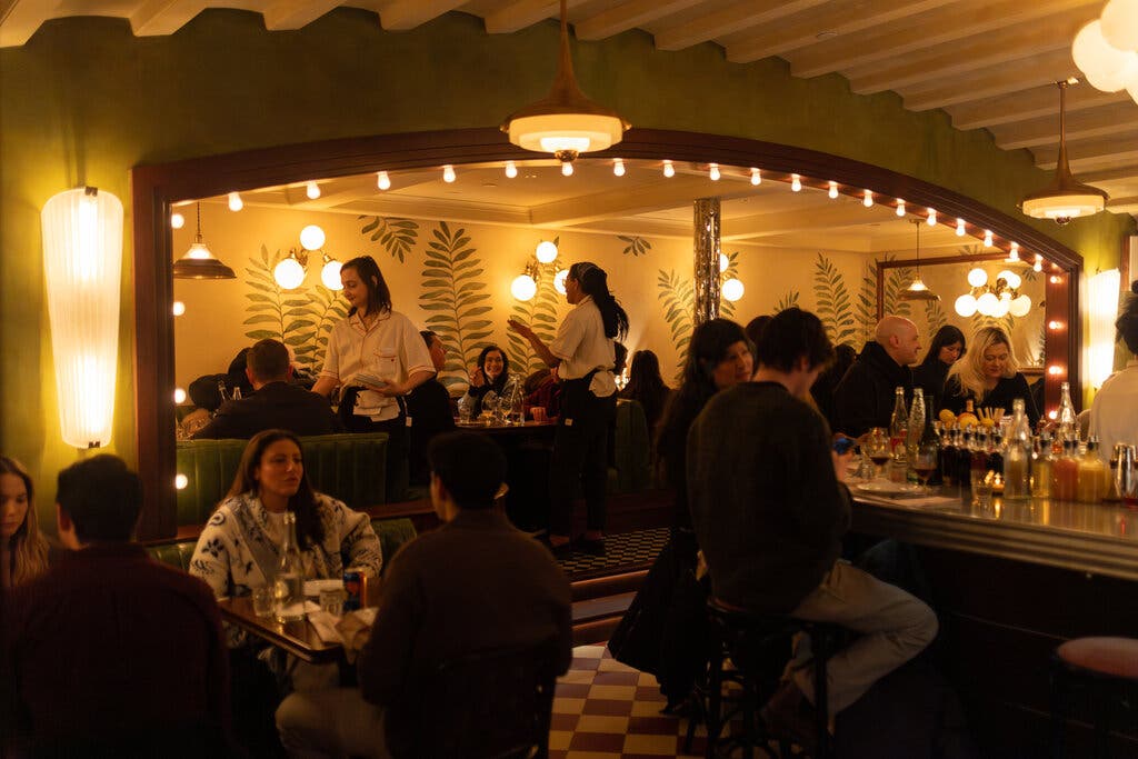 The interior of the restaurant, with numerous people seated at tables and a long zinc bar. The room features harlequin tile floors, lime green walls, a proscenium arch lined with bare bulbs, and wallpaper with fern designs.