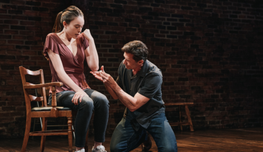 A man kneels on the floor, gesturing toward a seated woman who looks down with a serious expression; both are on a stage with brick walls and wooden chairs.
