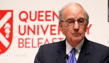 George Mitchell, with short grey hair, wearing glasses, a navy suit, white shirt and purple tie. He is standing behind a white podium. There is a white wall behind him with Queen's University Belfast and the university logo in red.