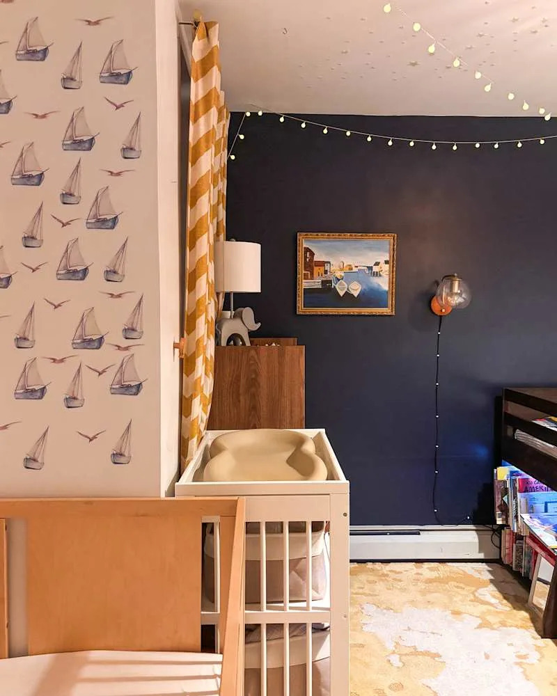 Nursery with sailboat wallpaper, yellow striped curtains, changing table, and a cozy rug, illuminated by string lights.