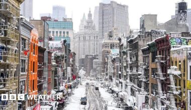 A snow covered main street lined with colourful buildings that have fire-escape stairs on the outside. In the distance are skyscrapers.