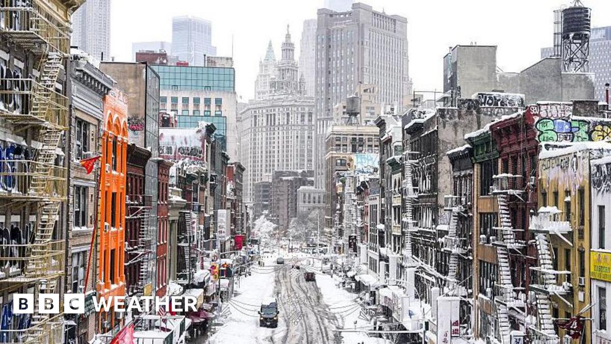 A snow covered main street lined with colourful buildings that have fire-escape stairs on the outside. In the distance are skyscrapers.