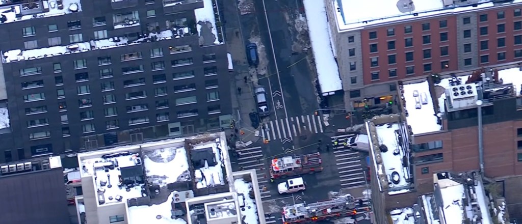 Aerial view of a snowy street with emergency vehicles and workers following a manhole explosion.