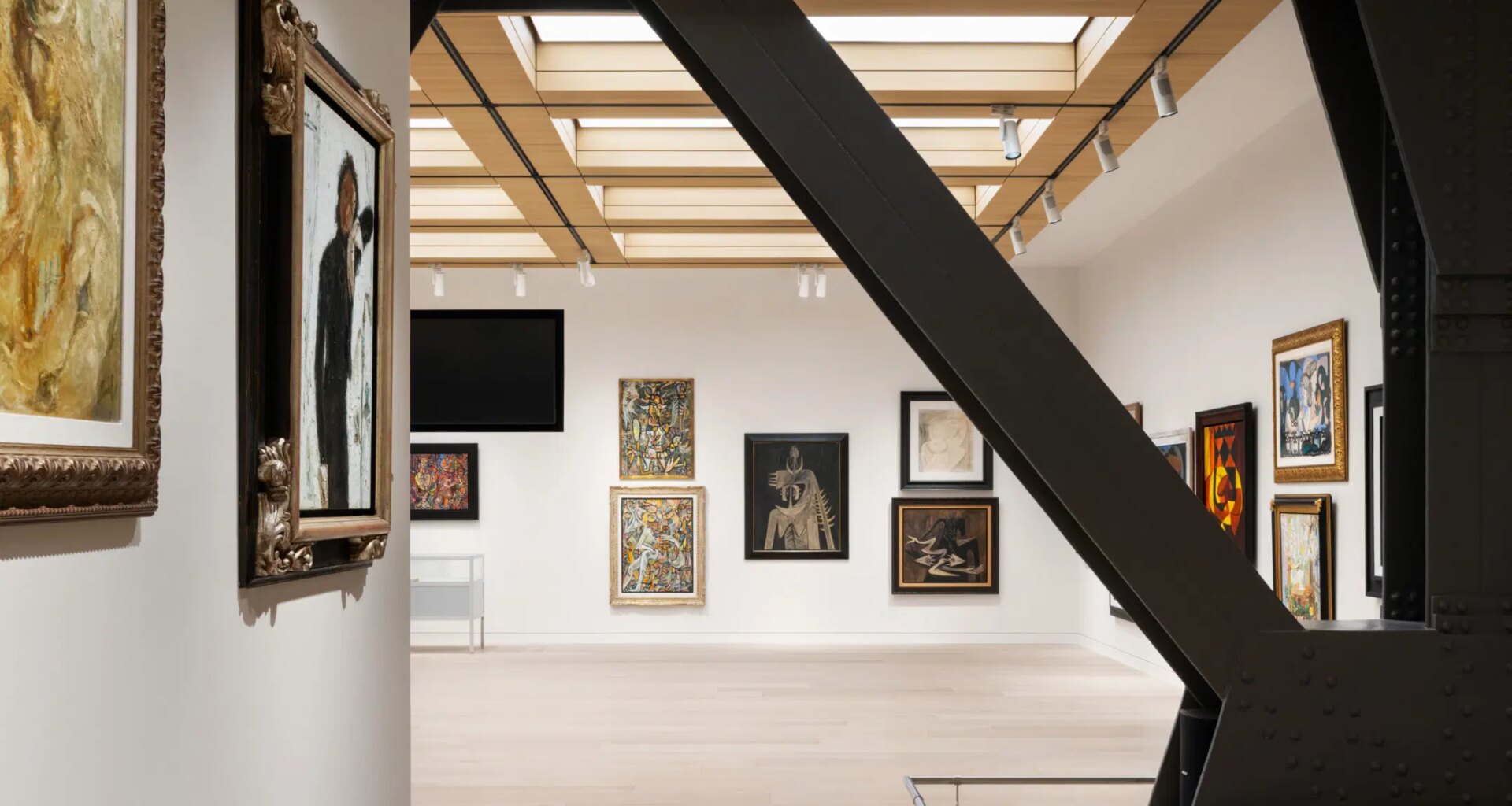 Modern art gallery interior with various framed paintings on white walls, featuring a large metal beam in the foreground.