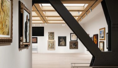 Modern art gallery interior with various framed paintings on white walls, featuring a large metal beam in the foreground.