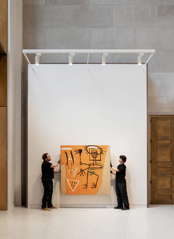 Two people hanging a modern abstract painting on a large white wall indoors.
