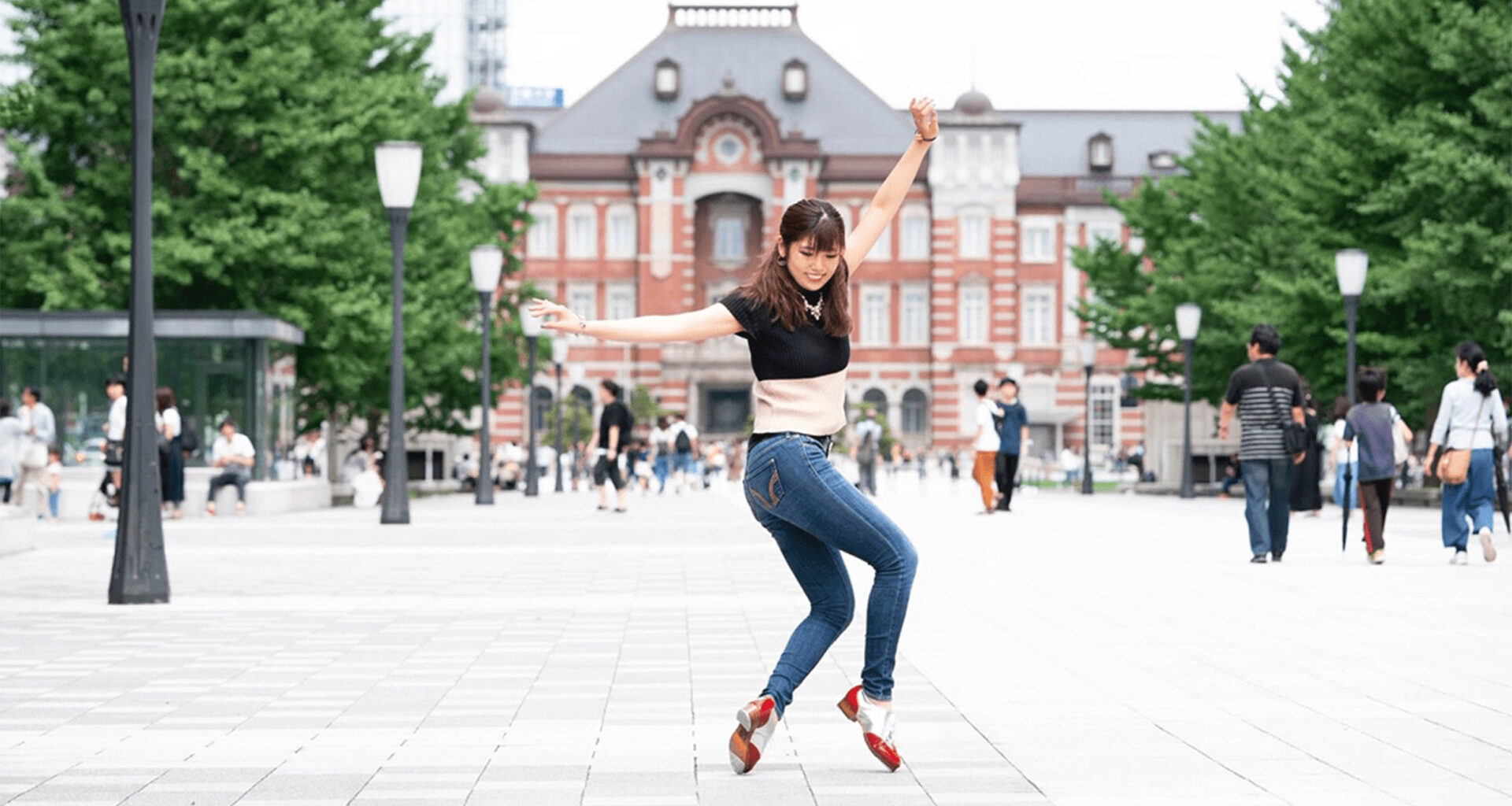 A woman in a black top and jeans performs a dynamic dance move on a plaza with a historic red brick building in the background. Other people are walking by and trees line the square.