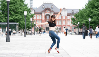 A woman in a black top and jeans performs a dynamic dance move on a plaza with a historic red brick building in the background. Other people are walking by and trees line the square.