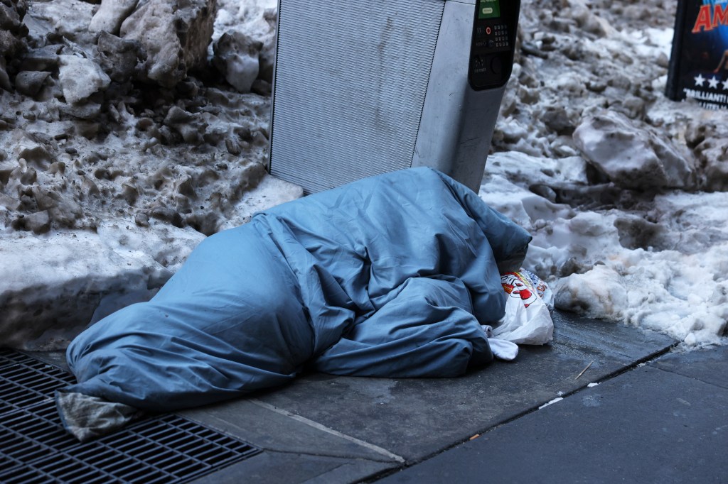 A person sleeping under a blue blanket on a snow-covered sidewalk next to a payphone.