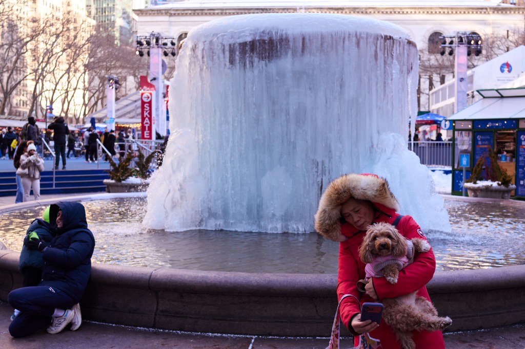 A woman in a red coat holding a small dog takes photos by a frozen fountain in Bryant Park, New York.